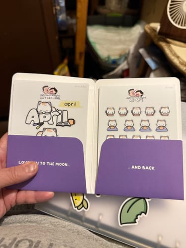 Customer holding the Cozy Cat Journal Pack open, showing two cute sticker sheets for 'April' inside purple pockets.