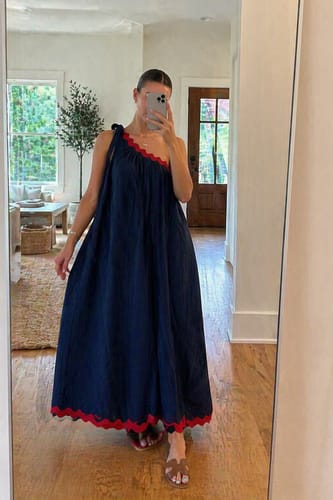 Reviewer in a navy one-shoulder maxi dress with a tied shoulder and red scalloped trim at the neckline and hem.