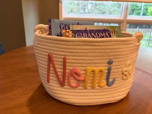 Customer's personalized white woven basket with the name 'Nomi' in colorful letters and a flower icon, filled with books.
