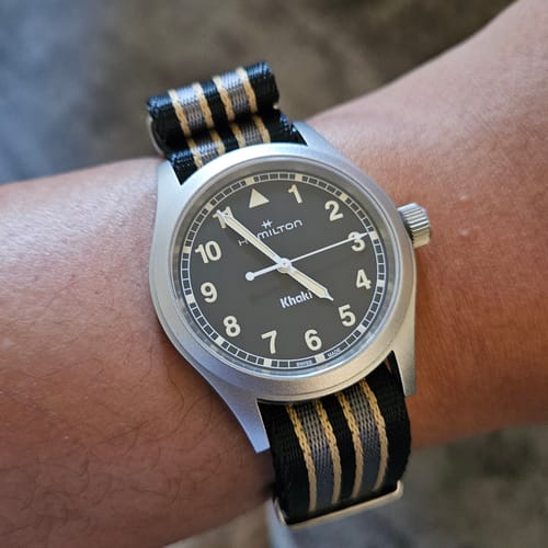 Reviewer's wrist showing the striped black, gray, and yellow AlphaPremier - Bond 2021 strap on a watch.