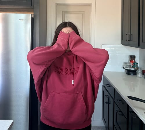Customer wearing the oversized red BTS Arirang hoodie, covering their face with the sleeves in a kitchen.