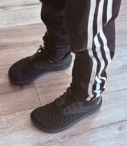 Customer's black Kid's Go I - Barefoot Shoes styled with black tracksuit pants on a wood floor.