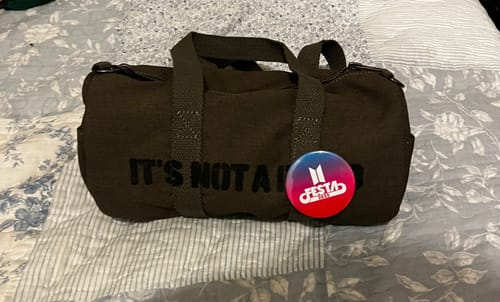 Reviewer's dark brown BTS Light Stick Bag with a colorful 'Festa' pin attached, sitting on a bed.