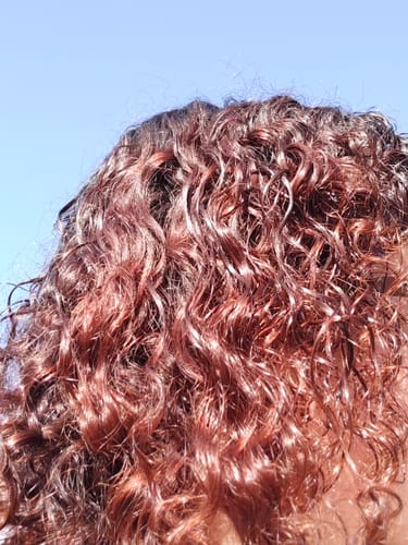 Clienta muestra el resultado del Tinte Castaño: cabello rizado brillante con reflejos cobrizos al sol.