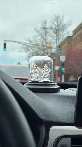 Reviewer's Silver Ganapathi & Lakshmi Idol in its glass dome, placed on a car dashboard.