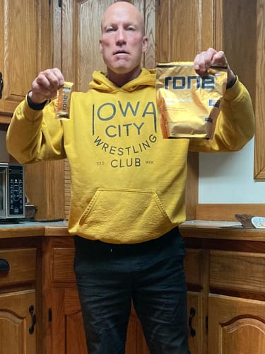 Reviewer in a yellow hoodie holding a single serving packet and a 20-pack of SuperCharged Honey in a kitchen.