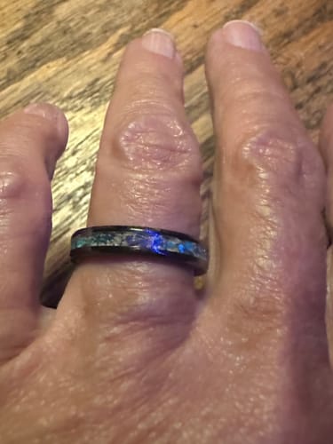 A customer's hand wearing a black carbide ring with an inlay of blue opal and cremation ashes.