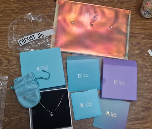 Customer's BTS X Stonehenge Coexist necklace in its box, with a blue pouch, several sleeves, and an iridescent bag.