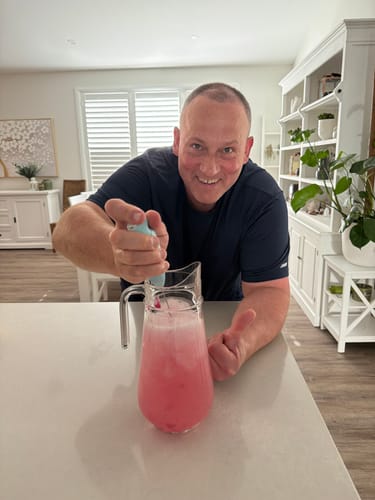 Customer happily mixing a pitcher of pink Original Enhanced Electrolytes drink in a kitchen, giving a thumbs up.