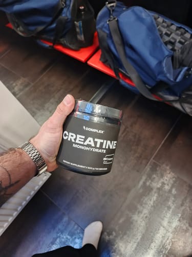 Reviewer's hand holding a black tub of Creatine Monohydrate in what appears to be a gym locker room.