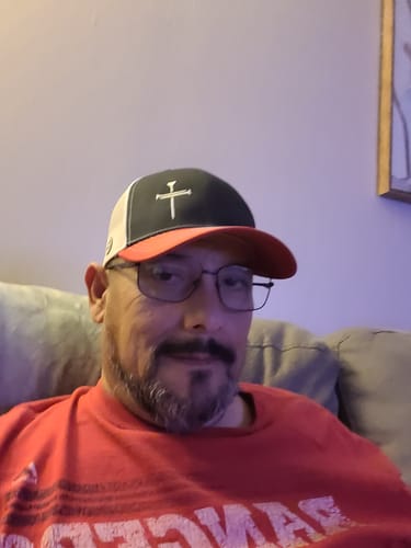Reviewer wearing the black, red, and white Nail Cross Everyday Faith Hat while seated on a couch.