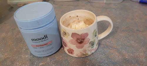 Customer's blue container of PM Blend next to a floral mug of hot chocolate with cream.