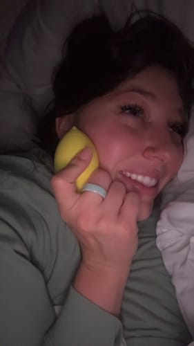 Reviewer smiling in bed while holding the yellow Lem Clitoral Massager to their cheek.