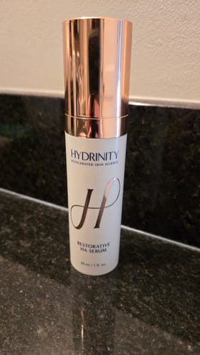 Reviewer's bottle of Hydranity Restorative HA Serum, with a white body and rose-gold cap, on a dark counter.