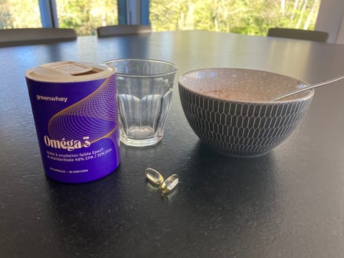 Client montrant son pot d'Oméga 3 et deux gélules sur une table de petit-déjeuner, à côté d'un bol.