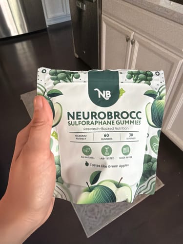 Reviewer holding the white and green pouch of NeuroBrocc Sulforaphane Gummies in their kitchen.