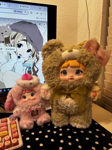 Customer's fluffy brown Nommi Good Night Bear plush with a green bow, next to a smaller pink one from the same series on a desk.
