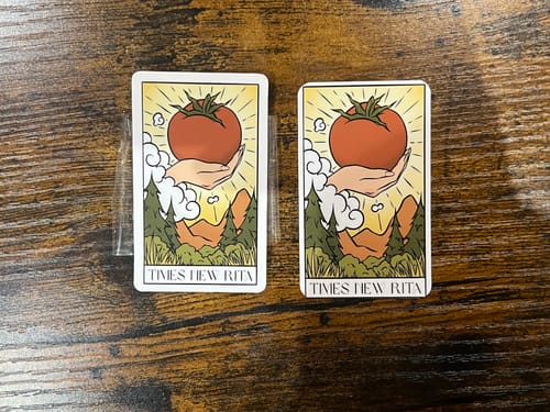Reviewer's two custom vinyl stickers on a wooden surface, showing a tarot card design with a hand holding a large tomato.