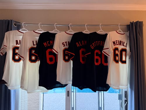 Customer displays a row of custom cream and black baseball jerseys with names and numbers on the back, hanging indoors.