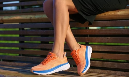 Reviewer sitting on a bench, wearing orange and blue Ortho Walk shoes with thick white soles.
