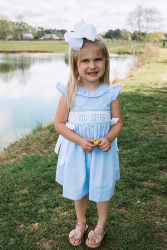 Reviewer's child smiling while wearing the Big Sister Blue Gingham Beverly Dress outdoors near a pond.