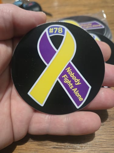 Customer holding a round, custom vinyl sticker with a purple and yellow 'Nobody Fights Alone' ribbon design.