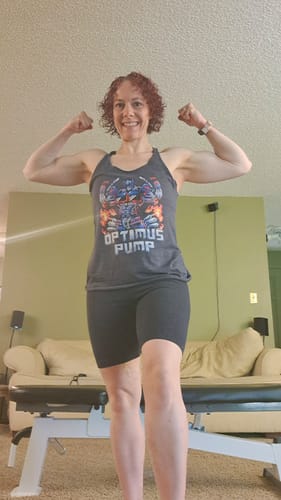 A customer flexes their arms while wearing the grey Optimus Pump Women's Racerback Tank in a living room.