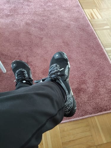 Arvostelijan jalassa mustat Vaistoa Lento -paljasjalkajuoksukengät sisätiloissa puulattialla.