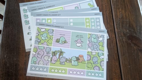 Reviewer's colorful Happy Dinos Planner Kit sticker sheets fanned out on a wooden table.