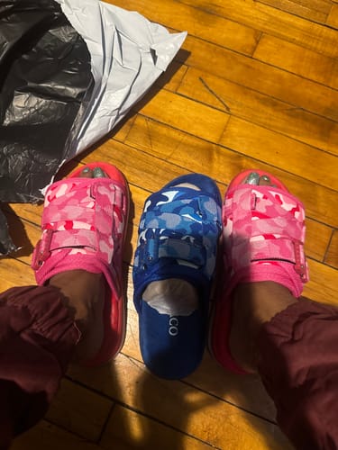 Customer wearing one pink camo Mystery DracoSlide, showing the pair alongside another blue camo slide on a wood floor.