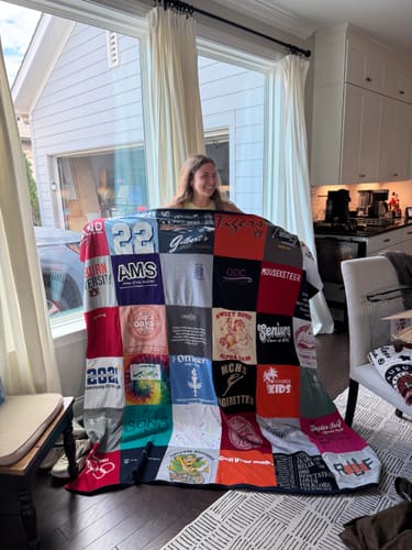 Customer smiling and holding up a large, colorful Project Repat T-Shirt Quilt in their living room.