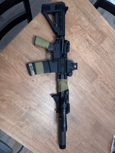 Reviewer's black rifle with OD Green Raid Tape wrapped on the grip, magazine, and handguard, shown on a wooden table.