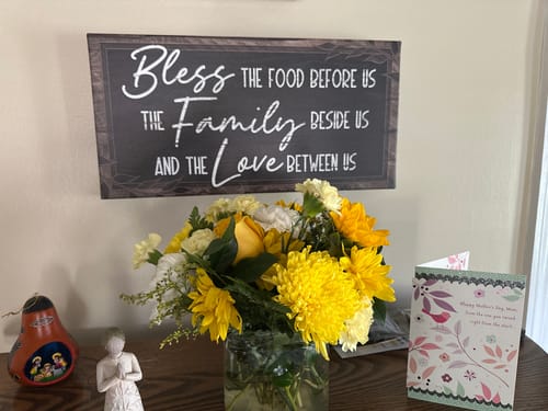Customer's 'Bless The Food Before Us' sign hanging on a dining room wall above a table with flowers and a card.