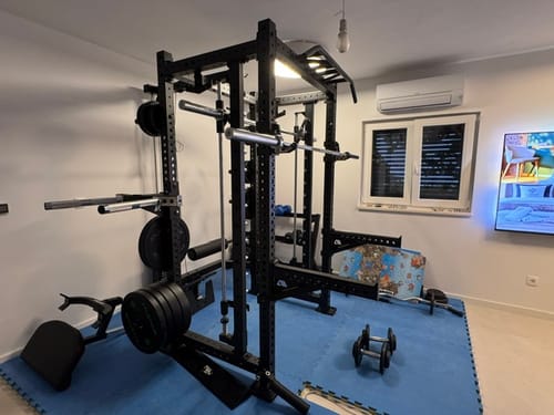 Ein Kunde zeigt sein schwarzes R8 Commander Power Rack von der Seite in seinem Heimstudio auf blauen Matten.