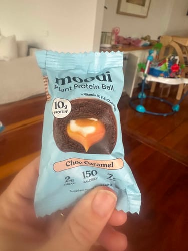 Customer holding the light blue package of a Choc Caramel Plant Protein Ball in their hand indoors.