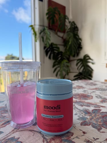 Customer's tub of Strawberry Watermelon Pre + Probiotic Blend on a table next to a glass of the prepared pink drink.