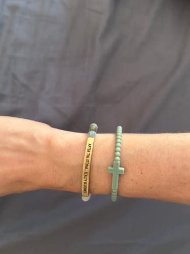 Customer's wrist showing the 'AFTER THE STORM, BEAUTY BLOOMED' engraved bracelet next to a cross bracelet.