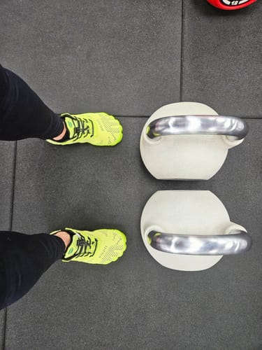 Reviewer standing in a gym wearing yellow Kid's Free Ⅰ - Barefoot Shoes next to kettlebells.