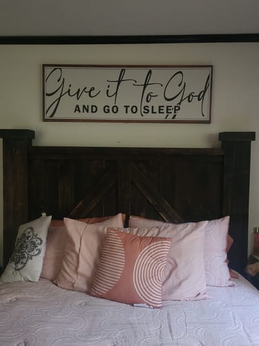 Customer's 'Give It To God And Go To Sleep' sign hanging in a bedroom above a dark wood headboard.