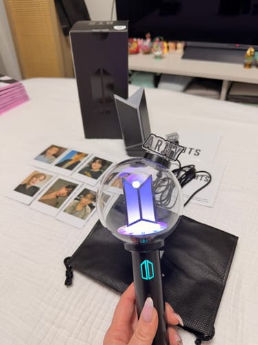 Reviewer holding the BTS Official Light Stick Ver.4, which is lit up in purple, with the box, cradle, and photocards nearby.