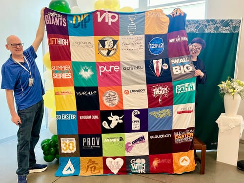 Two customers proudly holding up their large, colorful Project Repat T-Shirt Quilt made from many t-shirts.