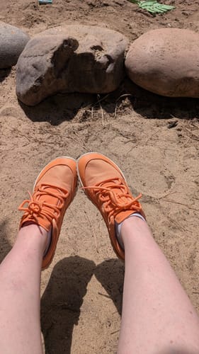 Reviewer wearing orange Forestep I barefoot shoes while sitting outdoors on the sand.