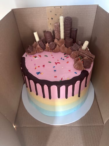 Customer's bespoke cake with rainbow-colored sides, pink icing, a chocolate drip, and assorted chocolate candy on top.