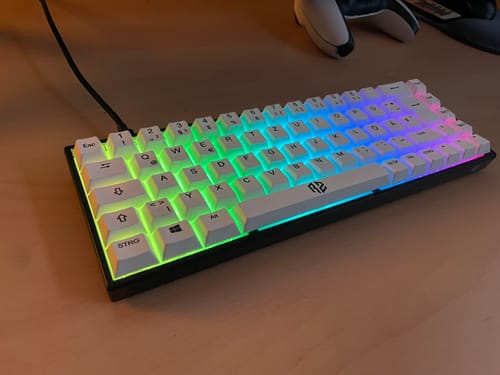 Rezensent zeigt die weiße RZ 65 HE Gaming-Tastatur auf einem Schreibtisch. Die Tasten leuchten in hellen, bunten Regenbogenfarben.