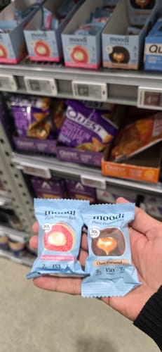 Customer's hand holding the Raspberry White Choc and Choc Caramel plant protein ball packages in a supermarket aisle.