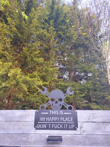 Reviewer's black 'This Is My Happy Place' metal wall art sign mounted on a fence outdoors in front of trees.