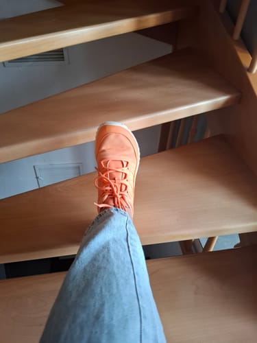 Ein Kunde trägt den leuchtend orangen Forestep Ⅰ Barfußschuh mit Jeans auf einer Holztreppe.