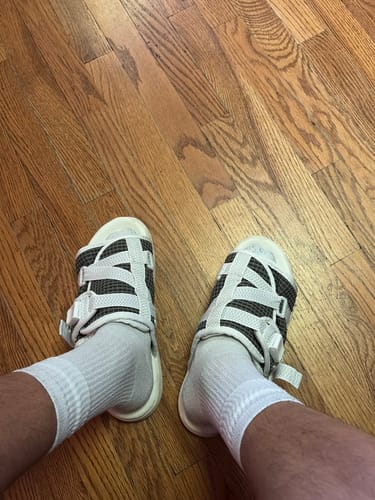 Reviewer's first-person view of their Mystery DracoSlides with a dark grid pattern and white straps, worn with white socks.