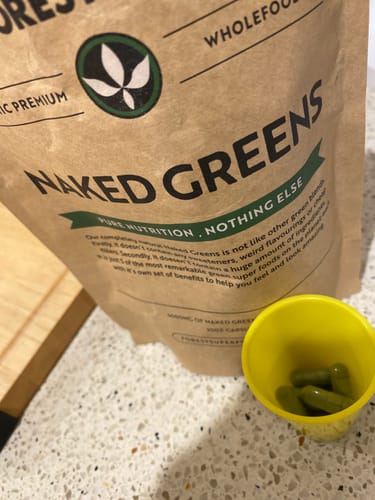 Reviewer's Naked Greens Capsules in a small yellow cup, with the brown product packaging behind it on a counter.
