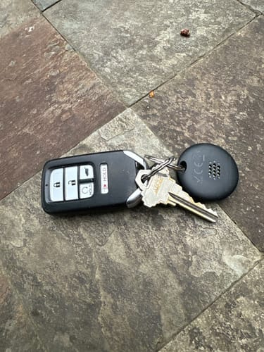 Customer's black SmartTag attached to a set of keys, including a car fob, lying on a stone floor.